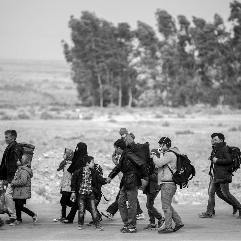 Age assessment of migrant children and young people A diverse group of people walking along a barren landscape, carrying bags.