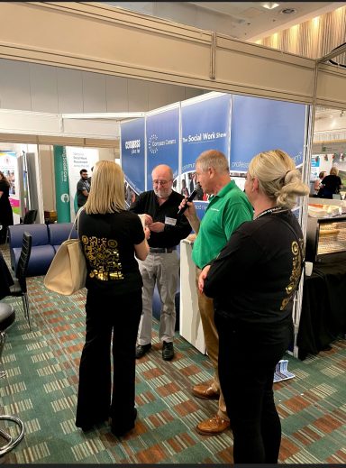 Neil Thomson picking K&C's raffle winner Three people conversing at a trade show booth, with one wearing a green sweater.