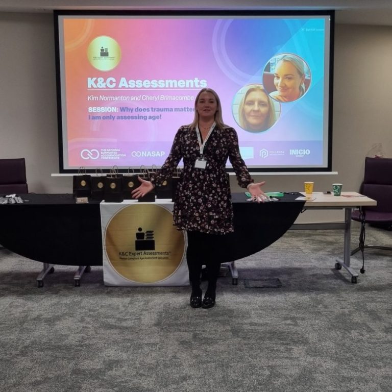 National Association of Supported Accommodation Conference 2024 Cheryl Brimacombe in a black dress stands before a presentation screen, arms outstretched, smiling.